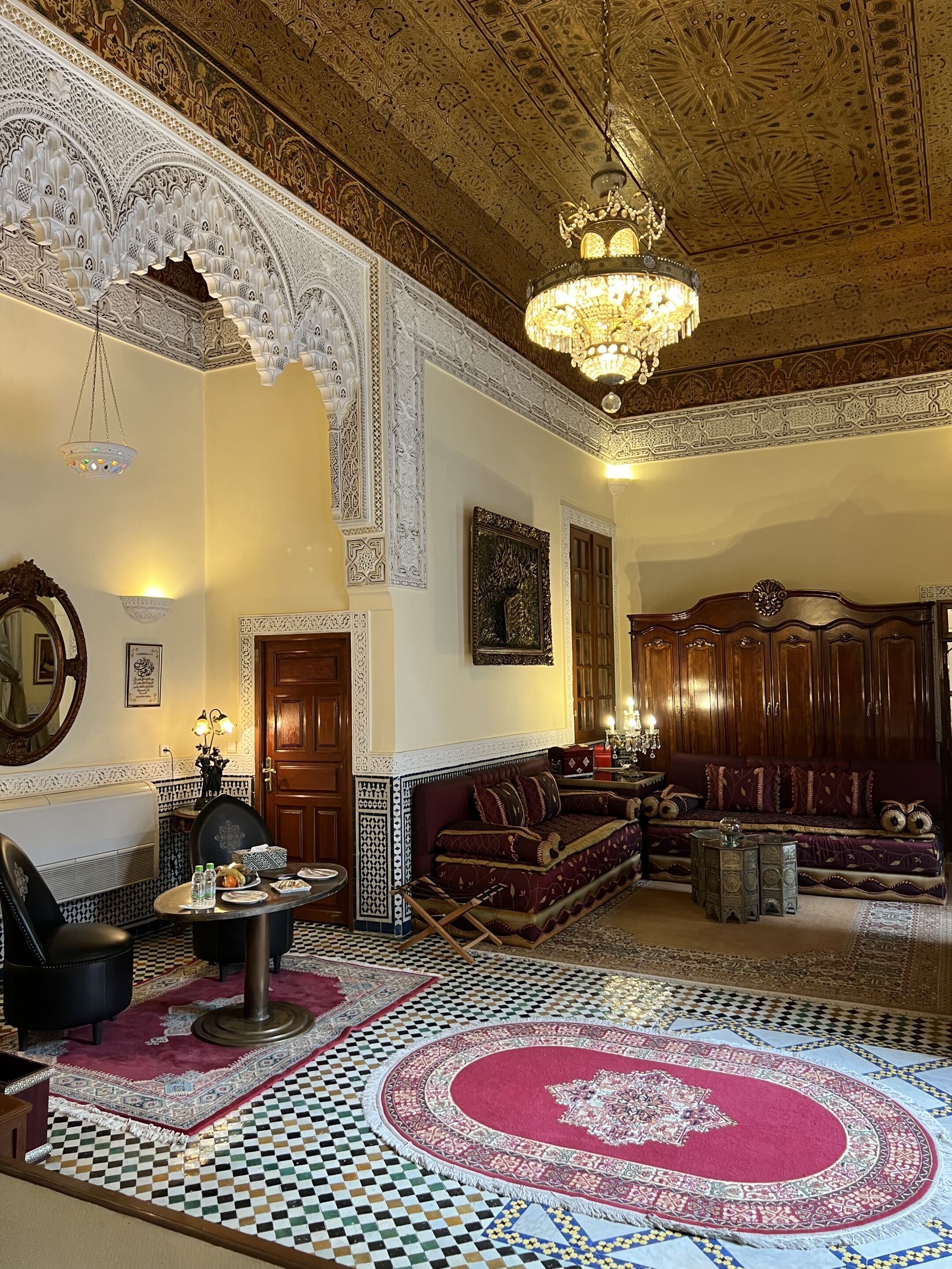 A richly decorated living room with intricate ceiling patterns, a chandelier, a sofa set, and a patterned floor.