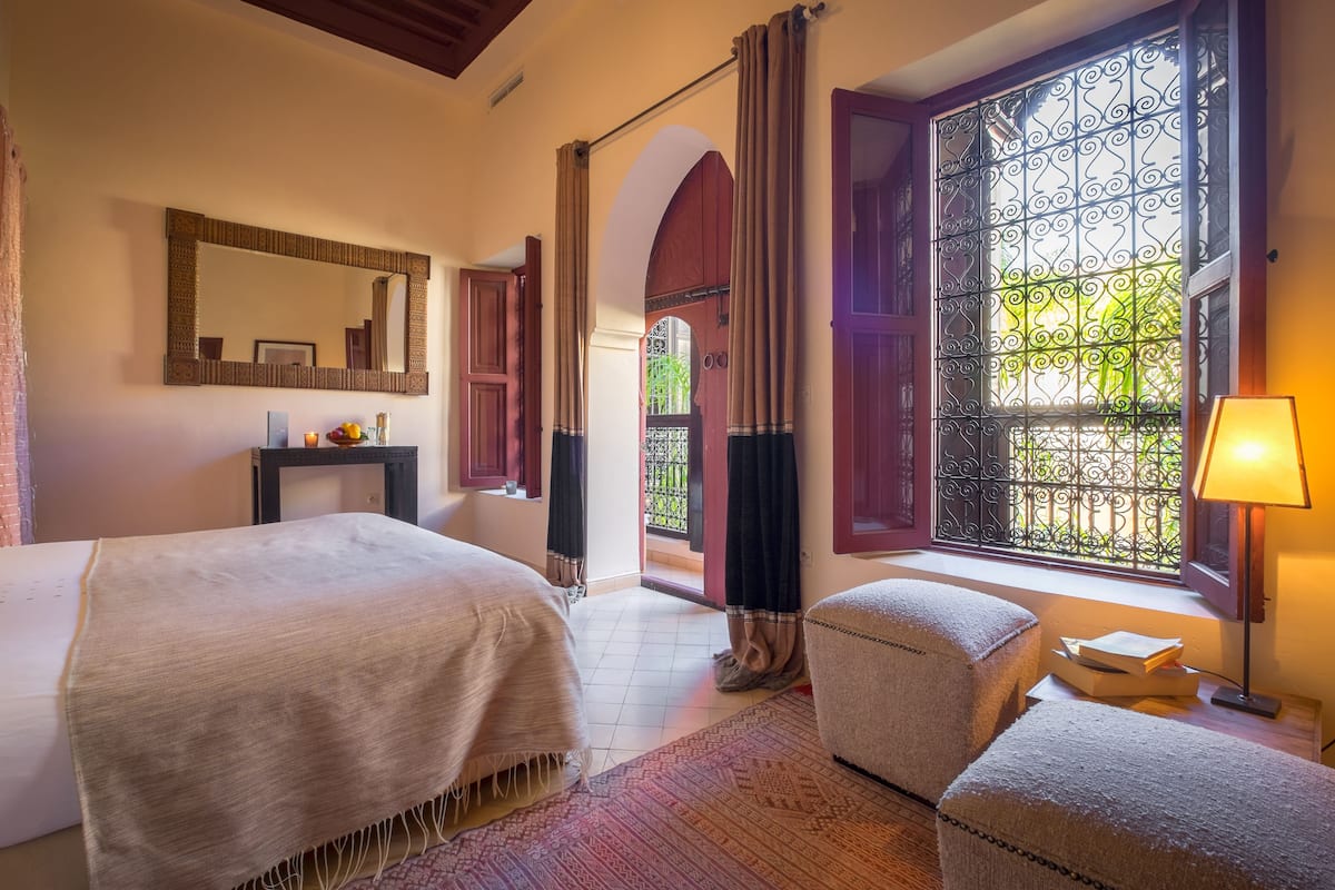 A hotel room with two beds, a small desk with a lamp, and a view through a window with decorative ironwork.
