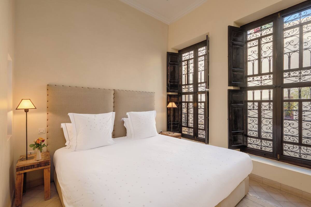 A neatly made bed with white linens, a bedside lamp, and a window with decorative iron bars.