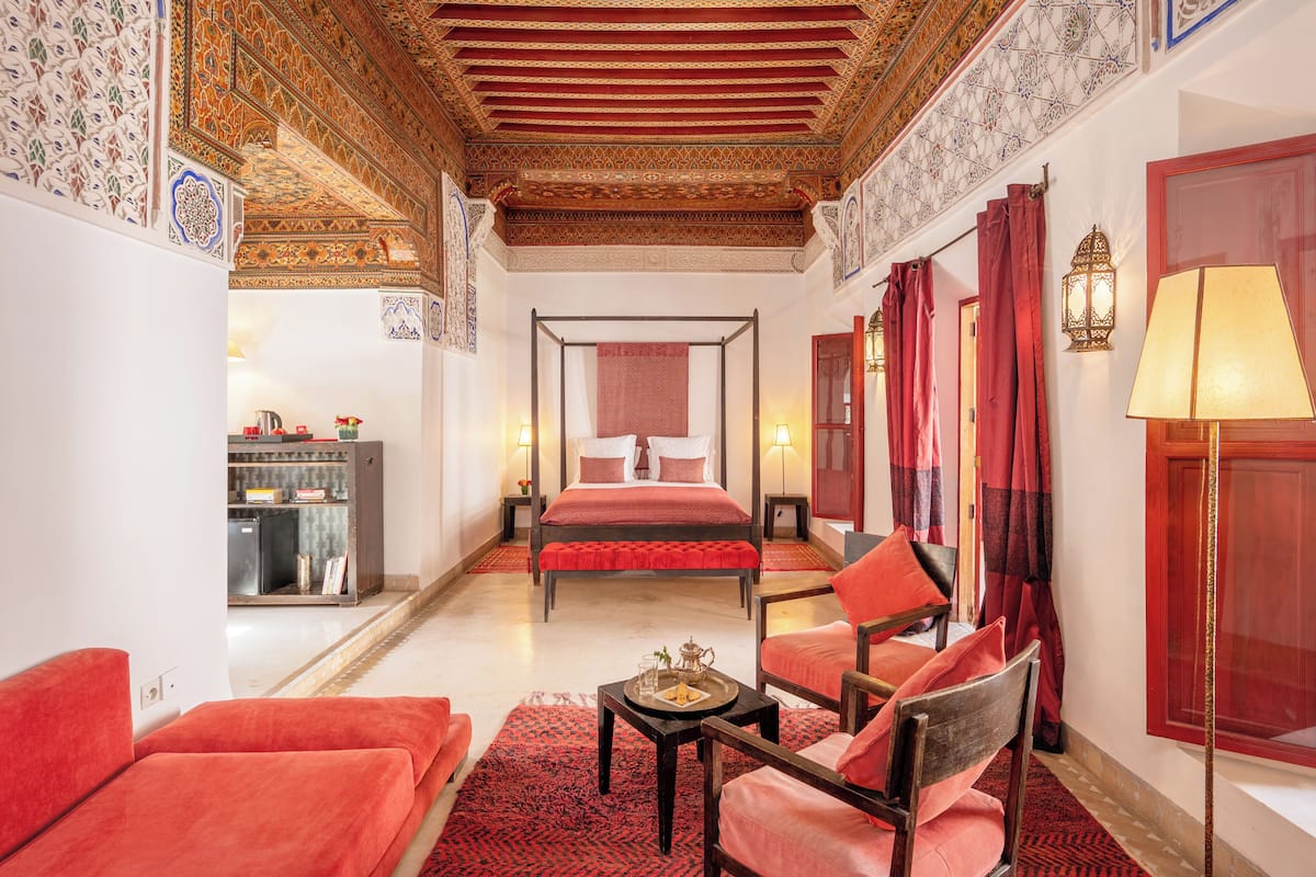 A traditional room with a four-poster bed, red upholstered chairs, a small table with a tray, and a decorative ceiling.