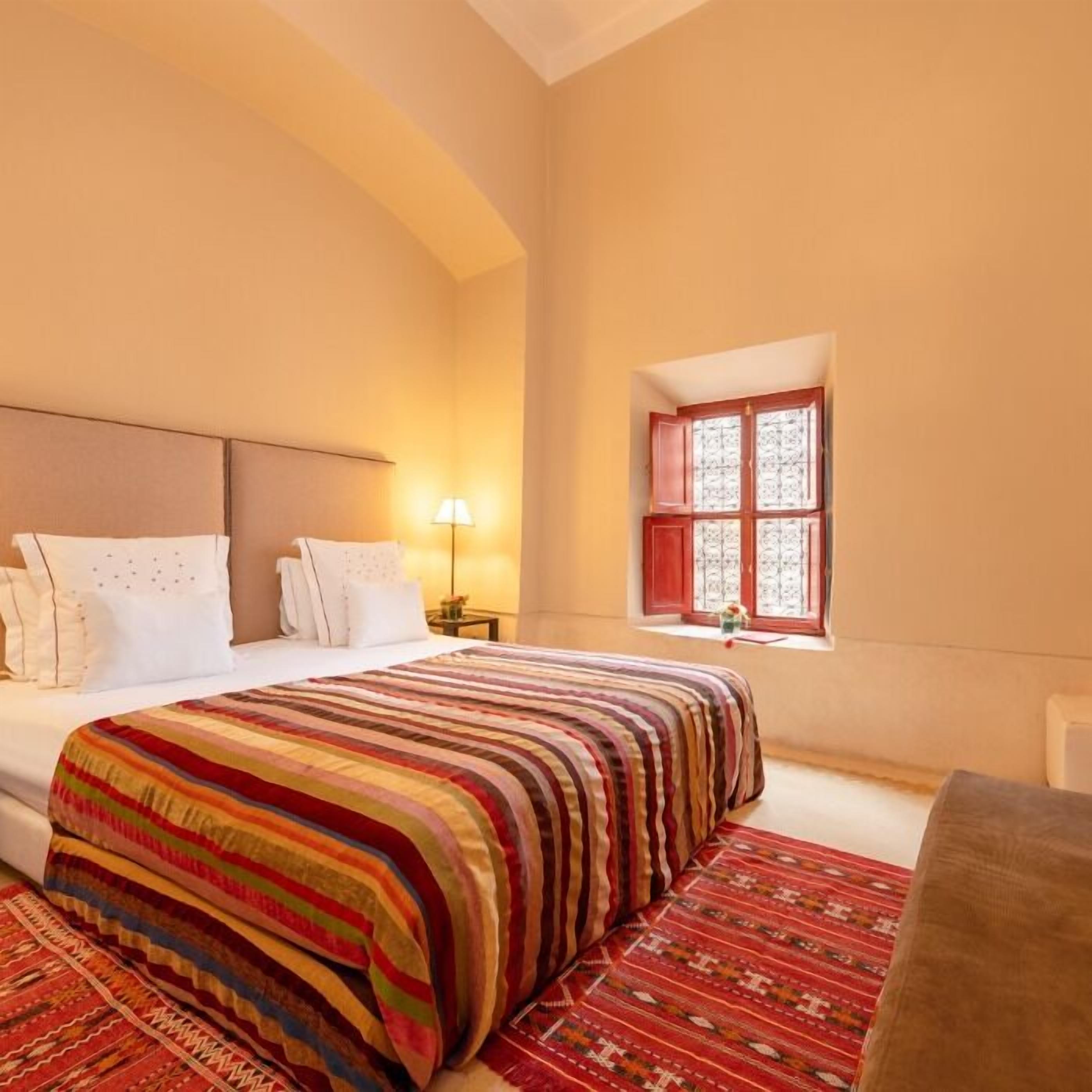 A bedroom with a large bed, a nightstand, a lamp, and a window with red shutters.