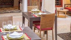 Un área de comedor al aire libre con mesas preparadas para dos personas, rodeada de cortinas blancas y un árbol.