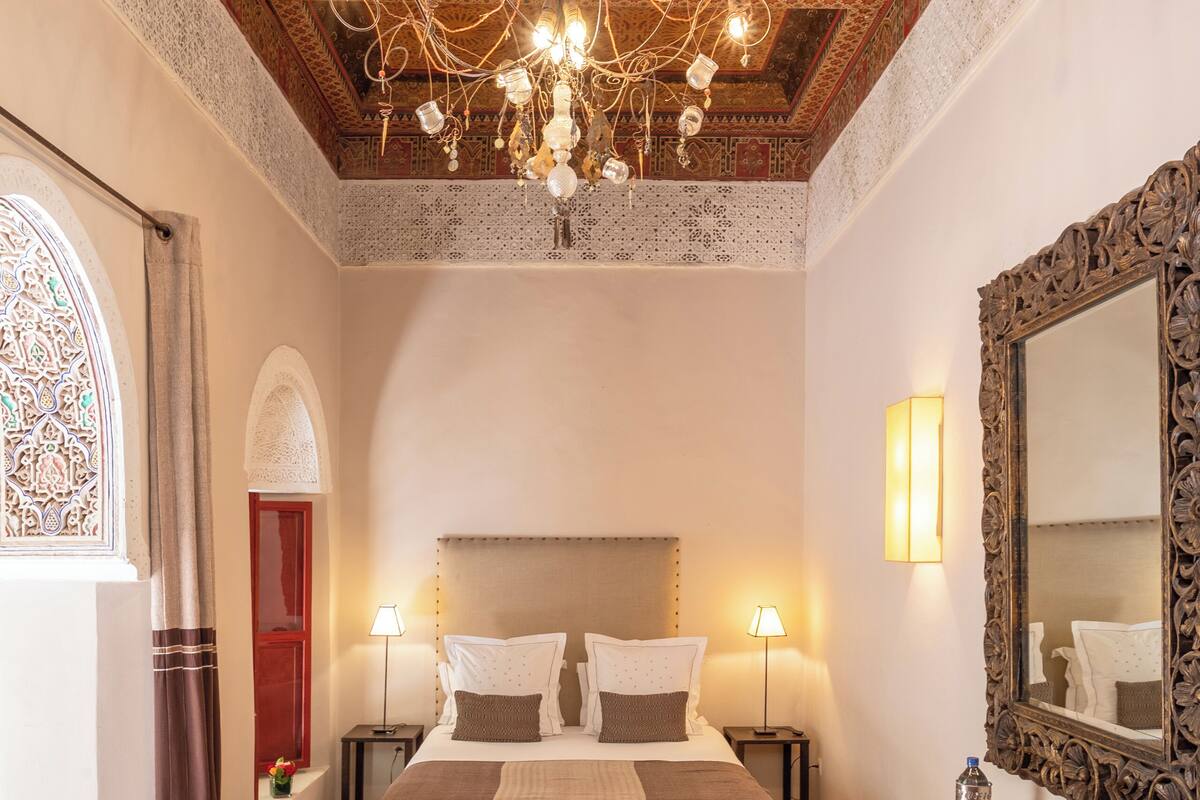 A bedroom with a large bed, a chandelier, a mirror, and a red bench.