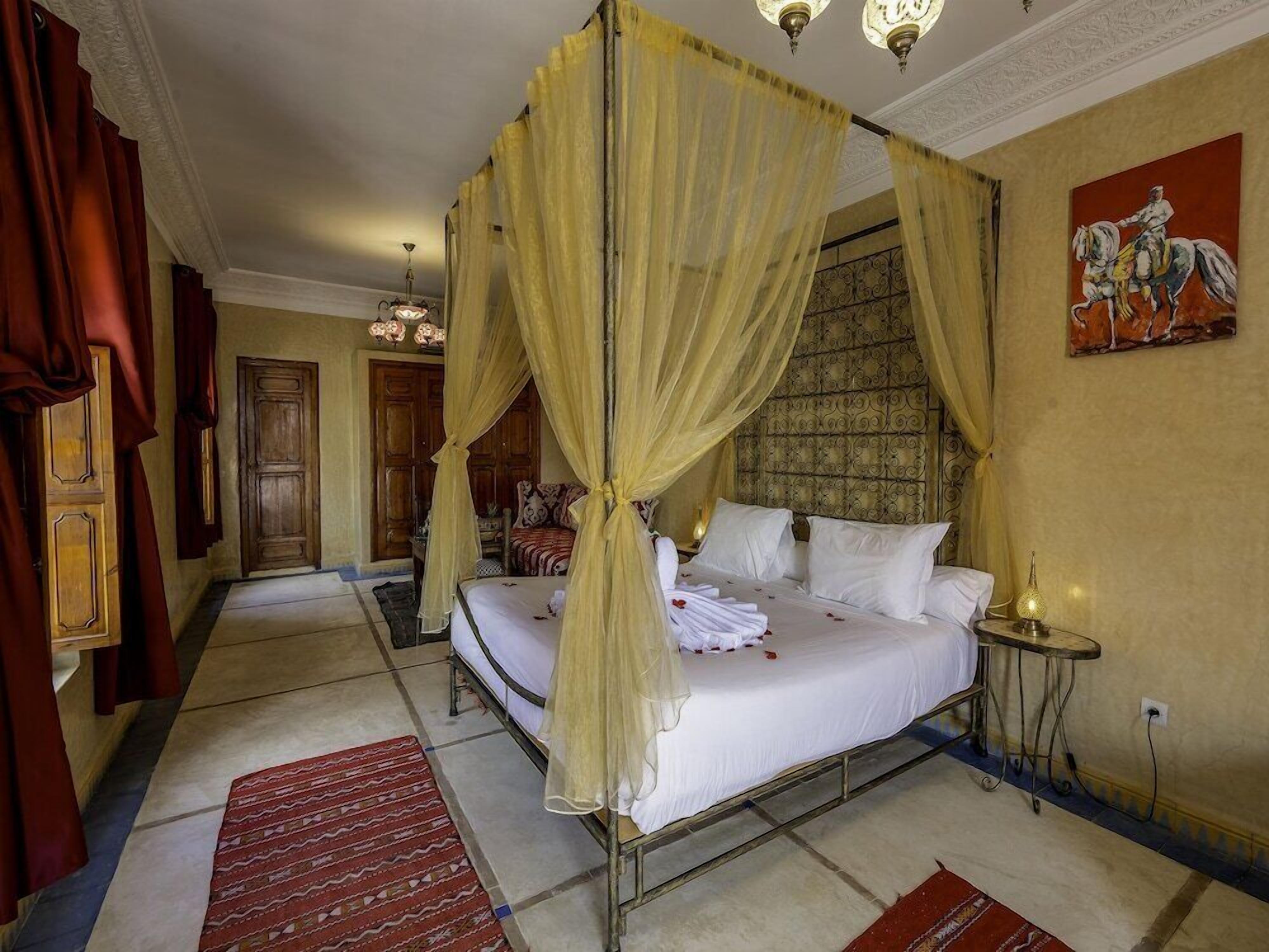 A four-poster bed with a canopy, a red patterned rug, a small table with a lamp, and a painting on the wall.