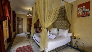 A four-poster bed with a canopy, a red patterned rug, a small table with a lamp, and a painting on the wall.