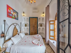 Une chambre d’hôtel avec un lit, une porte décorative, une étagère fixée au mur et une fenêtre avec un rideau.