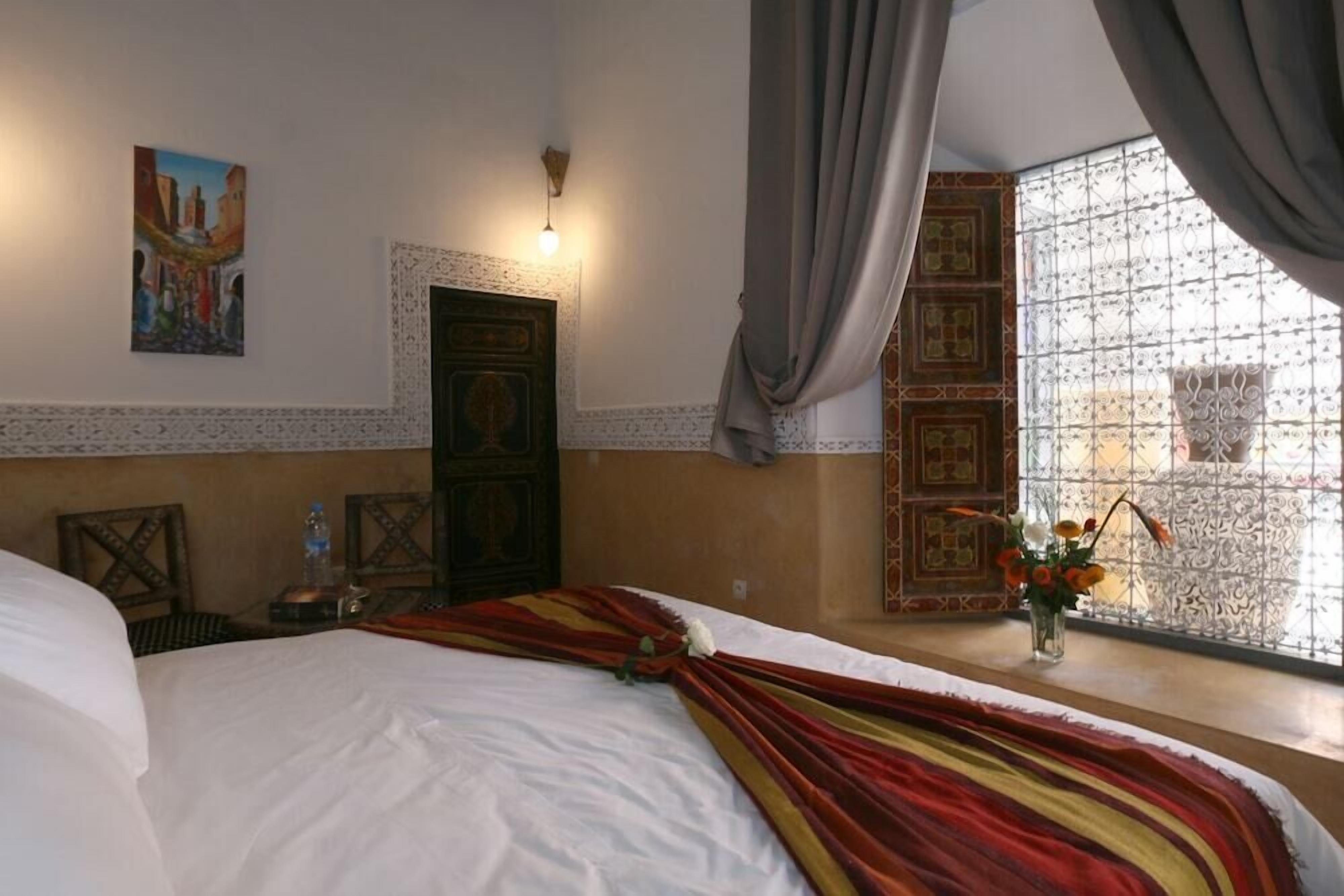 A hotel room with a bed, a painting on the wall, a door, and a window with curtains.