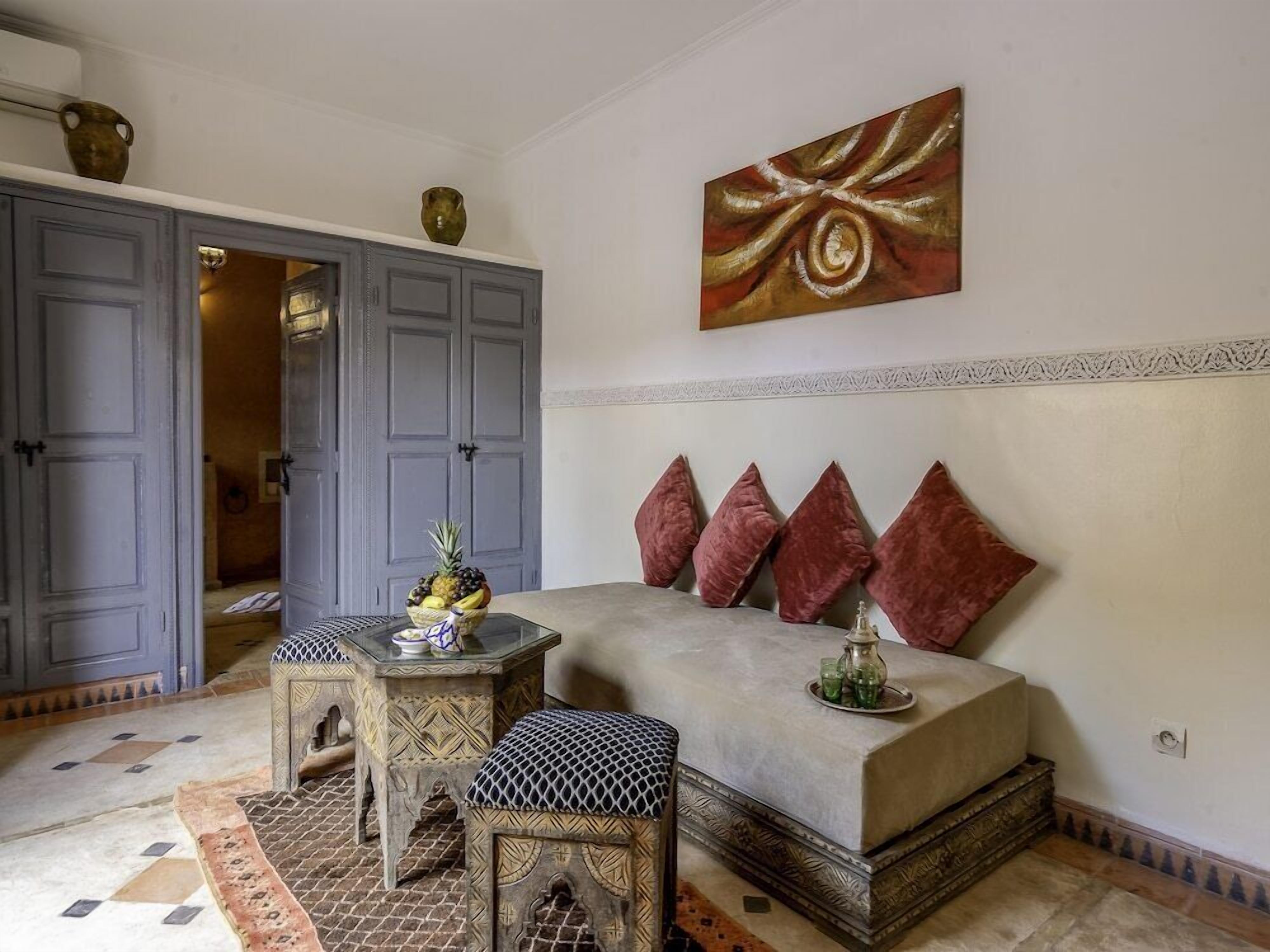 A room with a sofa, patterned ottoman, and a table with a fruit bowl.