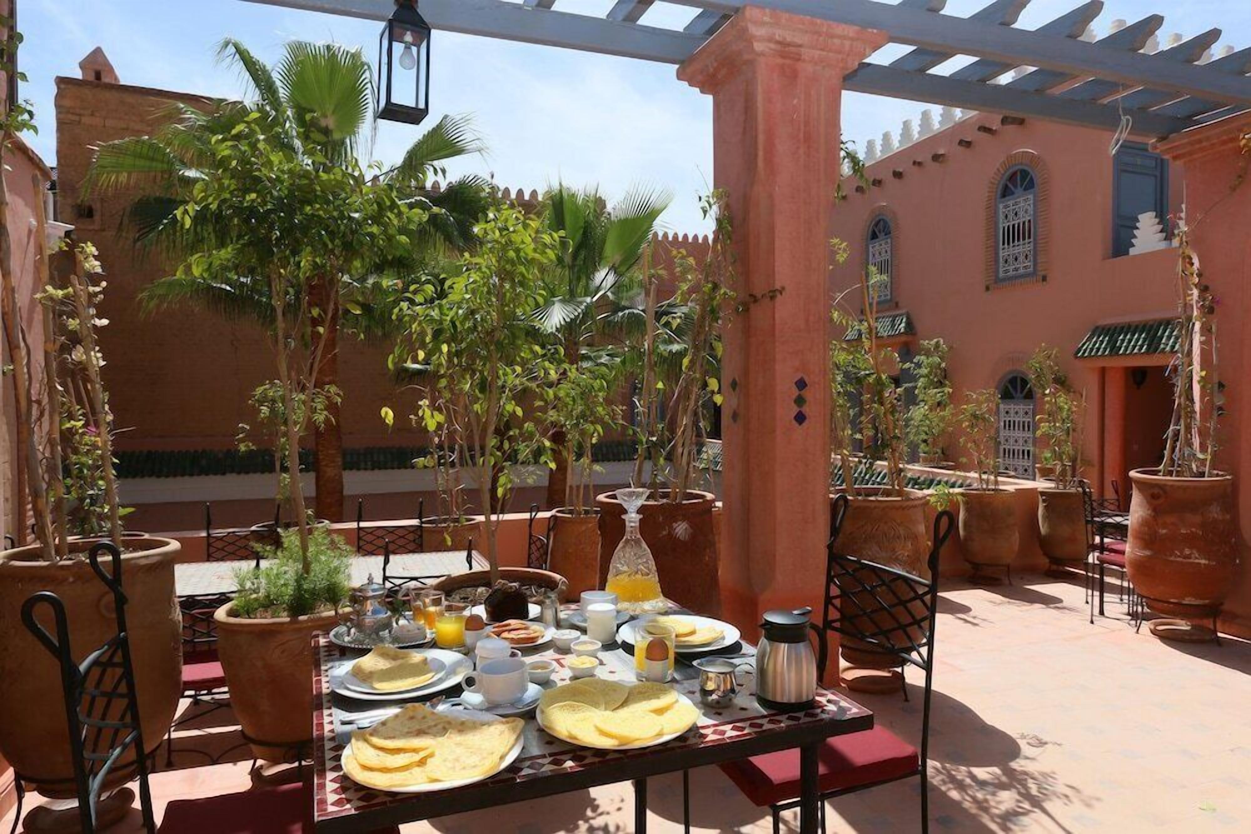 Un área de comedor al aire libre con una mesa lista para el desayuno, rodeada de plantas en macetas y un edificio de arquitectura tradicional.