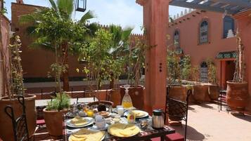 Un área de comedor al aire libre con una mesa lista para el desayuno, rodeada de plantas en macetas y un edificio de arquitectura tradicional.