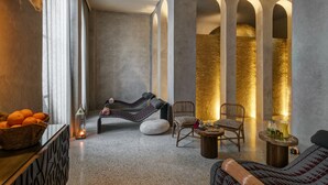 Een kamer in spa-stijl met een massagestoel, een houten schaal met mandarijnen, een klein tafeltje met een drankje en een gestructureerde muur met bogen.