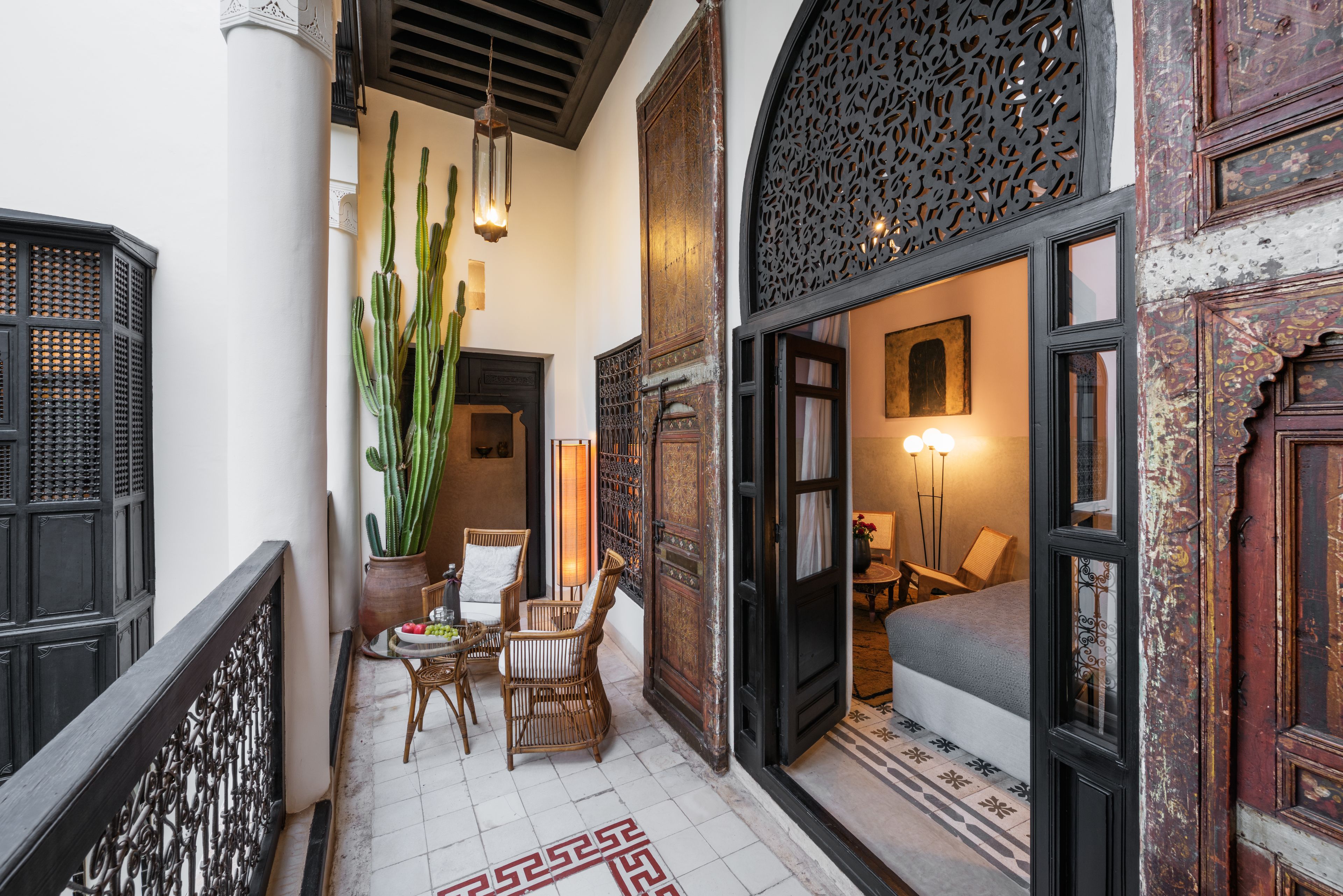 A narrow corridor with a wooden railing, a cactus plant, wicker chairs, and a patterned floor.