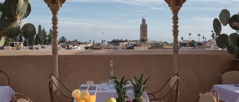 A terrace with a table set for breakfast, featuring fruit and drinks, overlooking a cityscape with a prominent tower.