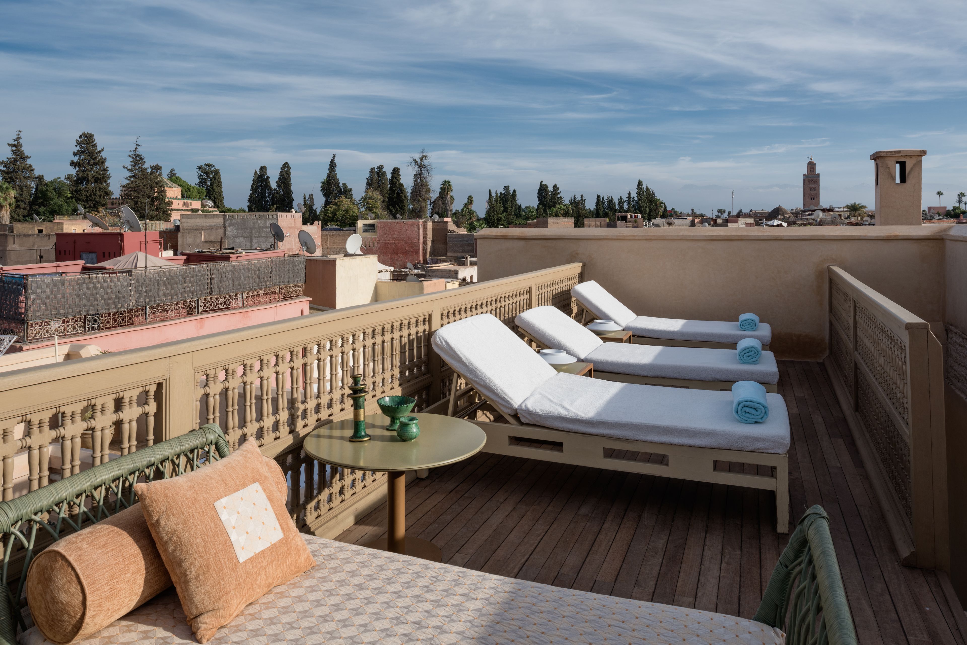 A rooftop terrace with lounge chairs and a table, overlooking a cityscape with buildings and a tower.