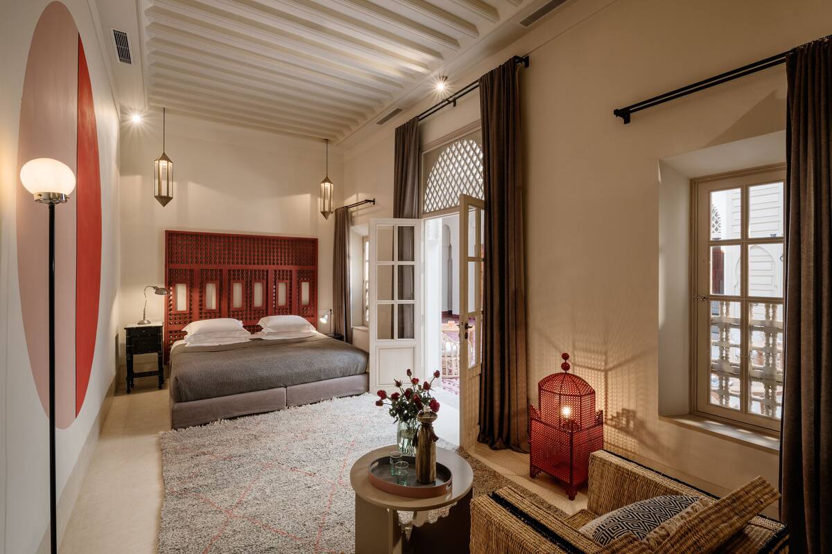 A bedroom with a large bed, a bedside table with a lamp, a small table with a vase and flowers, a red lantern, and a window with curtains.