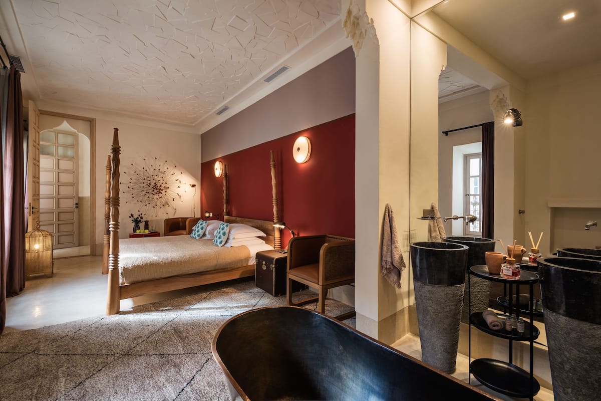 A bedroom with a large bed, a wooden chair, a bathtub, and a vanity with large black containers.