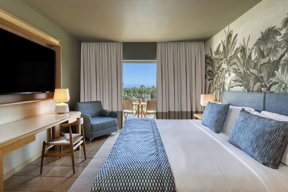 A hotel room with a bed, a desk, a TV, a view of the outdoors, and a dining area.