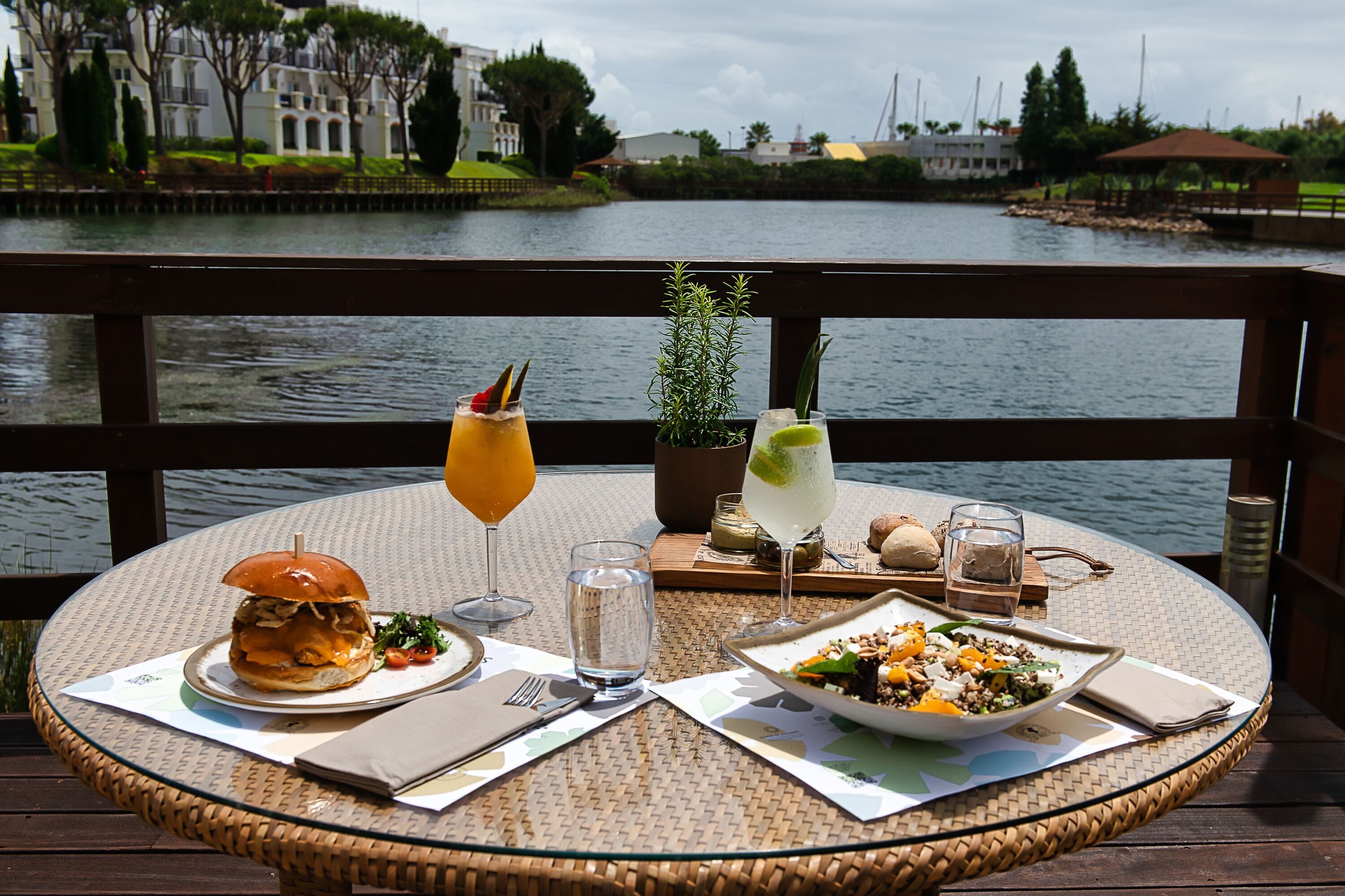 A dining table with a burger, salad, and a bowl of salad with fruit, accompanied by drinks and a small plant.
