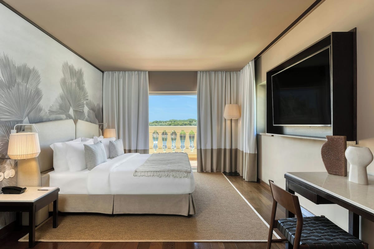 A modern hotel room with a large bed, a desk, a TV, and a balcony with a view of greenery.