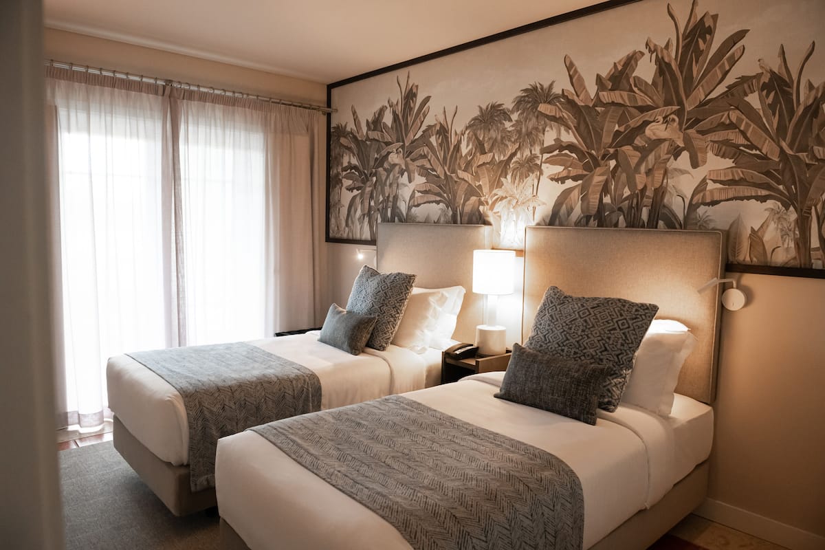 A hotel room with two beds, a large wall mural, a nightstand with a lamp, and a window with sheer curtains.