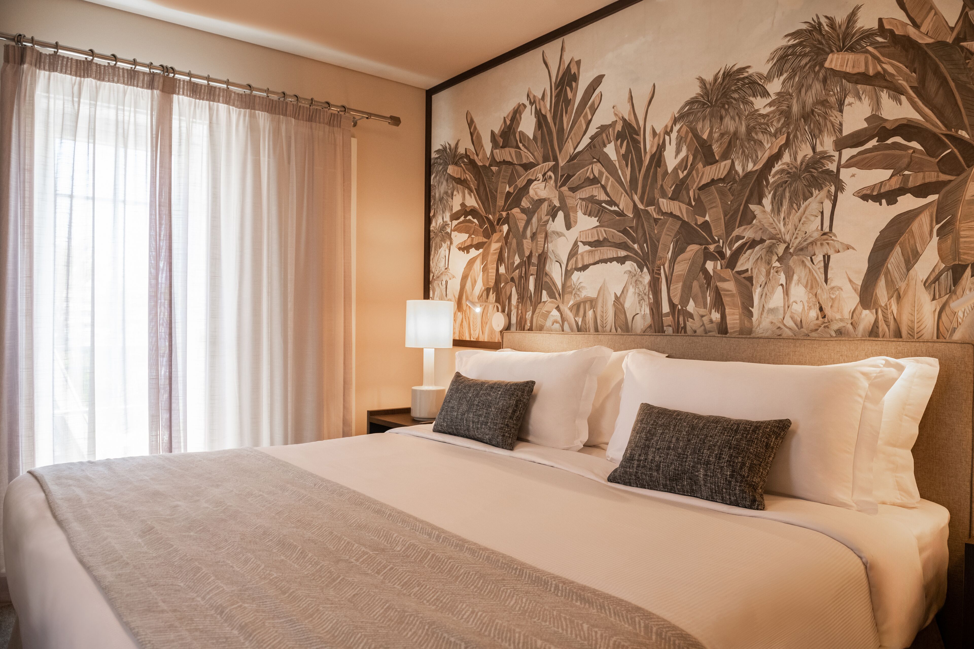 Habitación de hotel con una cama grande, una mesita de noche con lámpara y un mural con motivos tropicales en la pared.