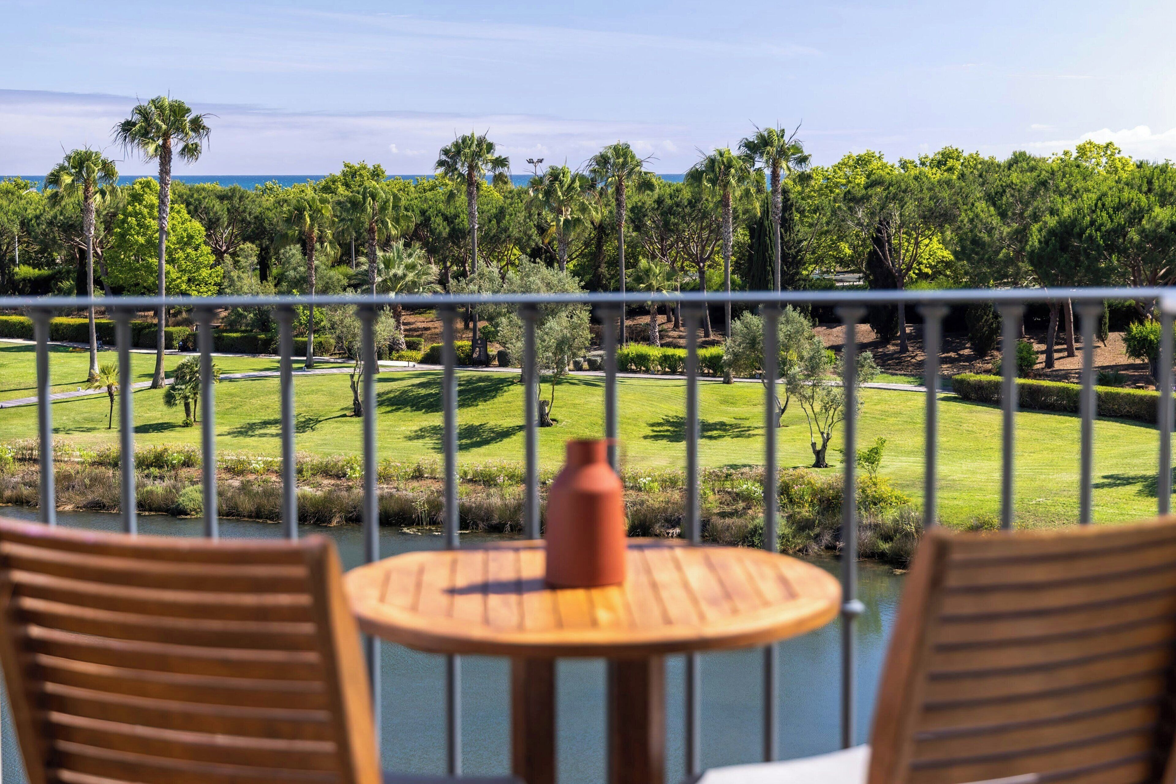 Un balcón con vista a un campo de golf, una pequeña mesa con un florero y dos sillas de madera.