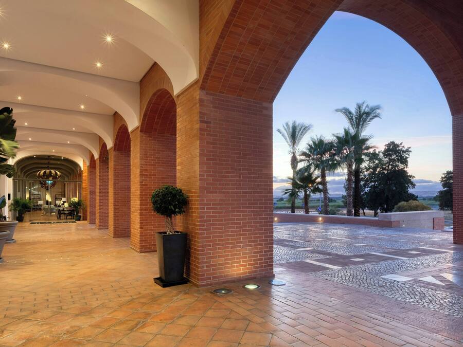 Un pasillo amplio con columnas de ladrillo y vistas a palmeras y un atardecer.