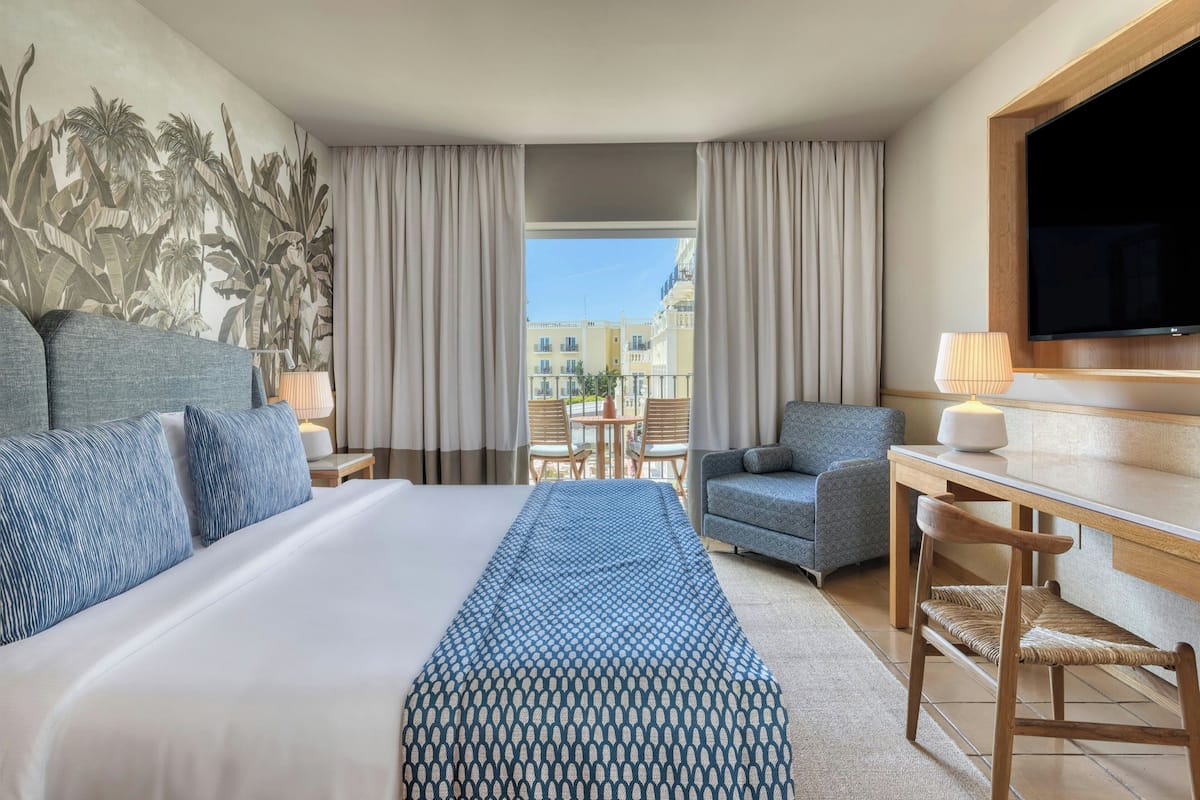 A hotel room with a large bed, a desk, a chair, a TV, and a balcony with curtains.