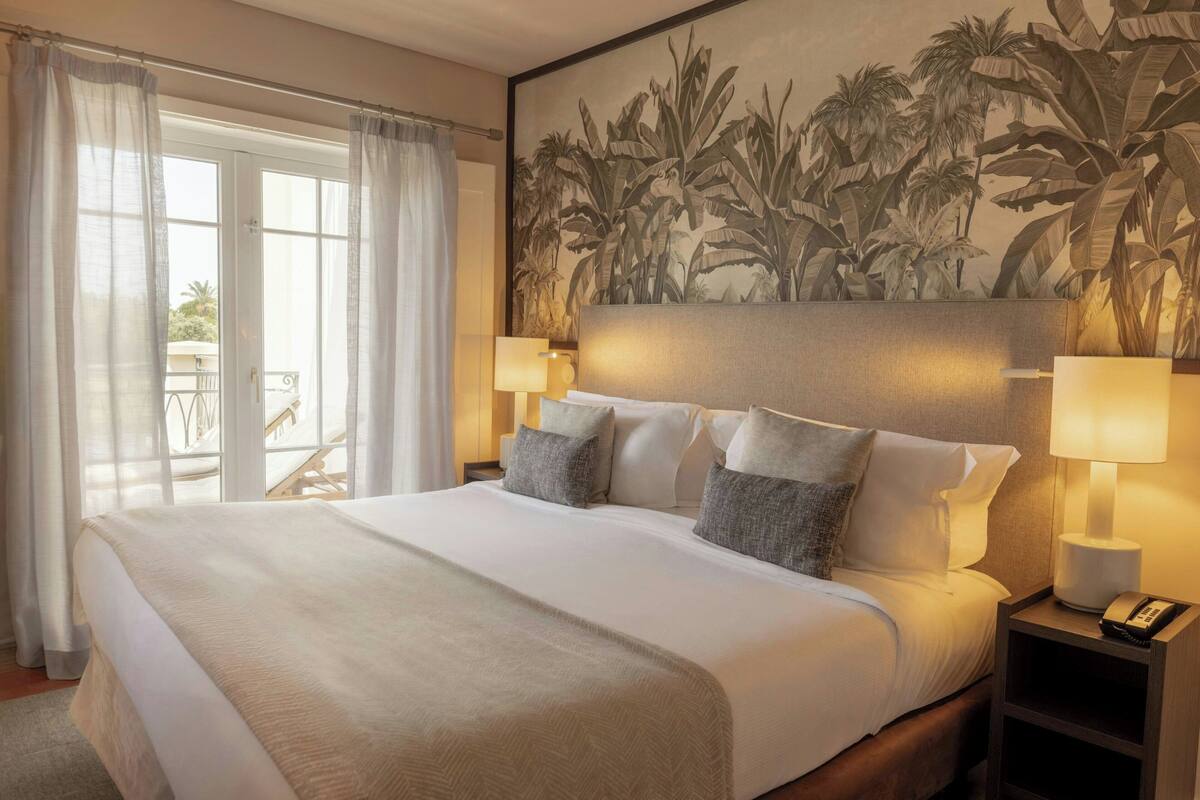 A hotel room with a large bed, two bedside lamps, a nightstand, a balcony with a chair, and a tropical-themed wall mural.