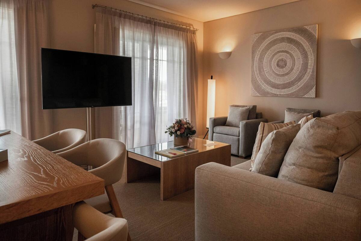A modern living room with a flat-screen TV, a sofa, a coffee table with a vase of flowers, and a wall-mounted artwork.