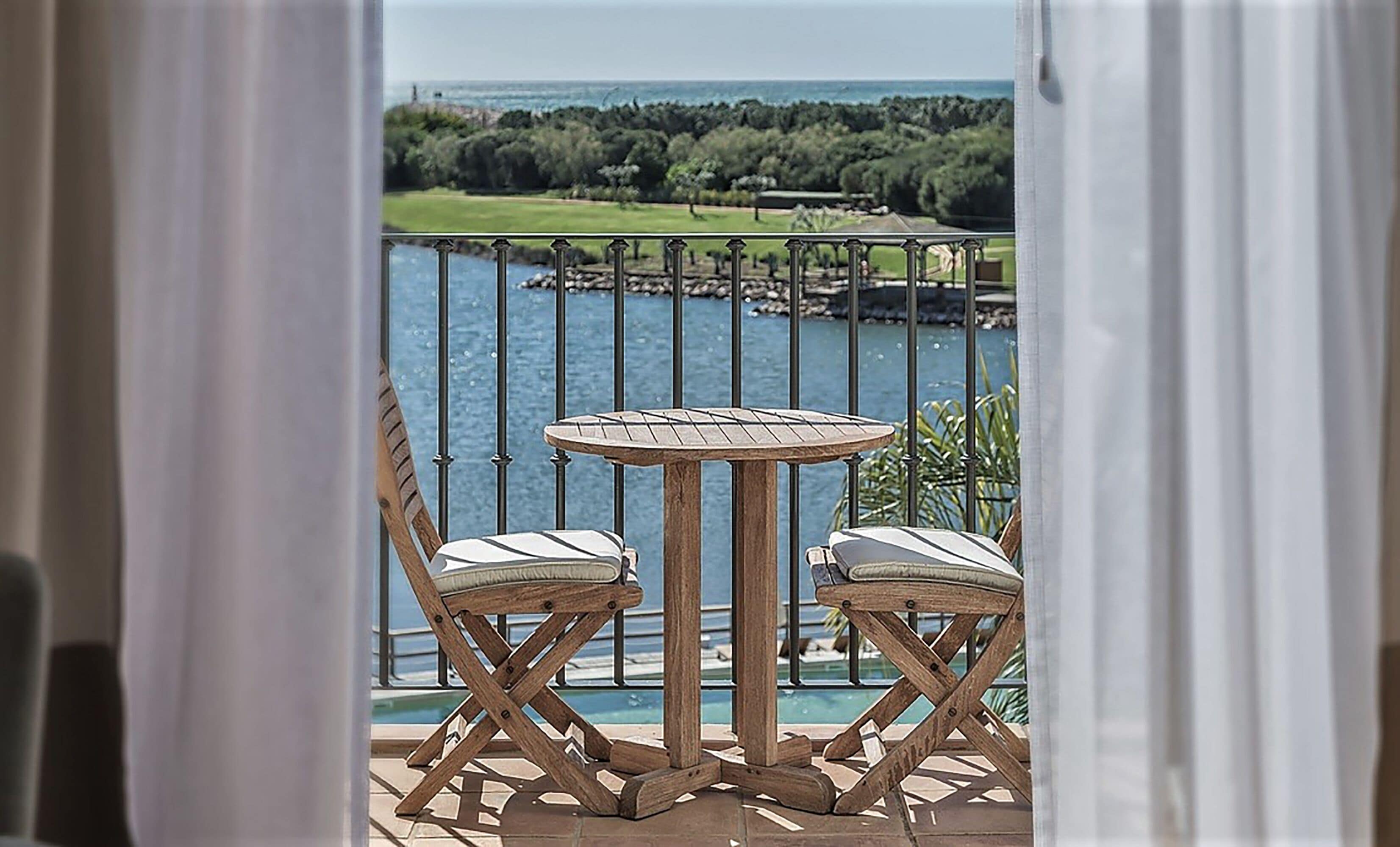 Un balcón con una mesa redonda de madera y dos sillas acolchadas, con vistas a un cuerpo de agua y un campo de golf.
