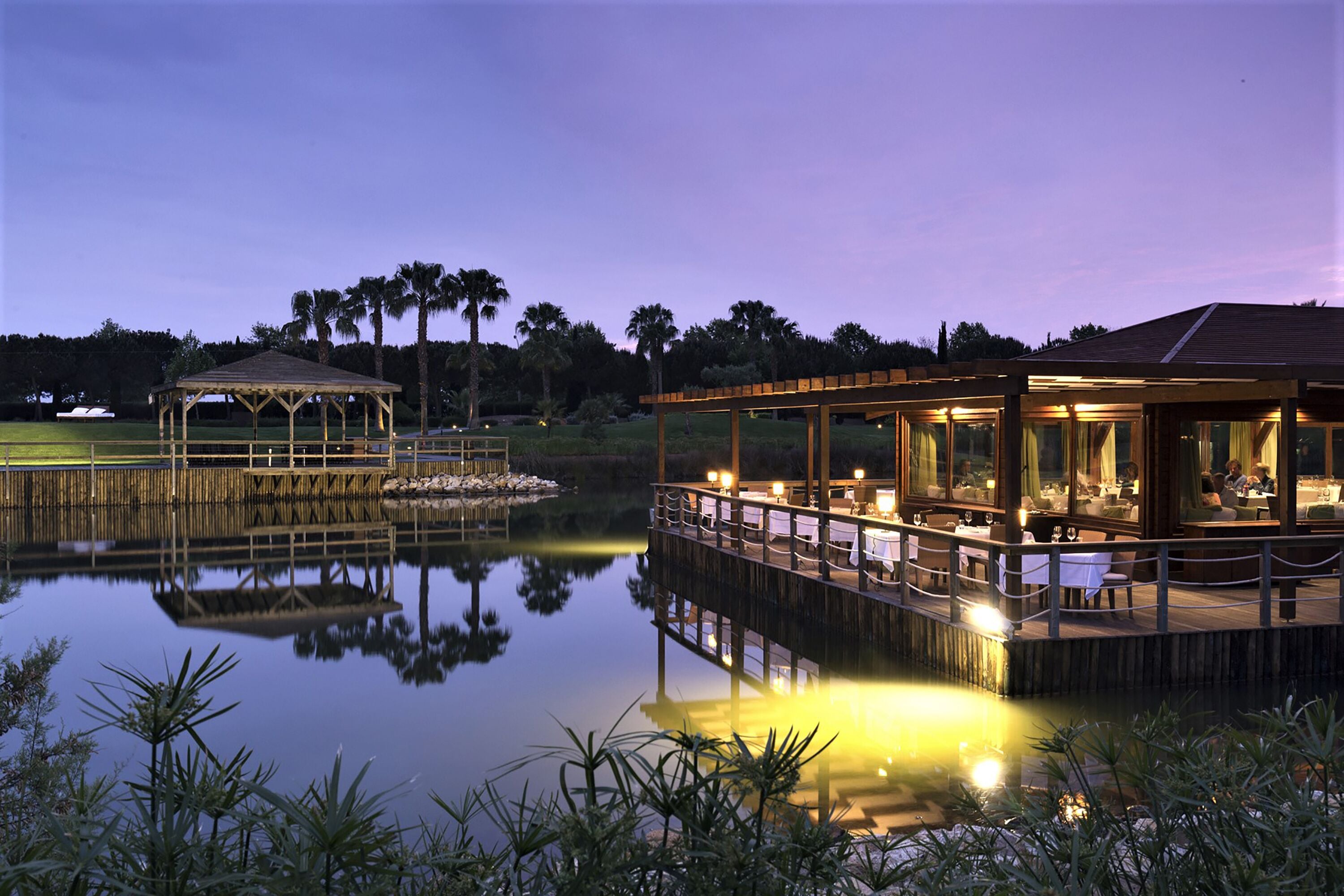 Un restaurante frente al agua con terraza al aire libre, vista a un lago tranquilo, un pabellón cubierto y una terraza de madera.