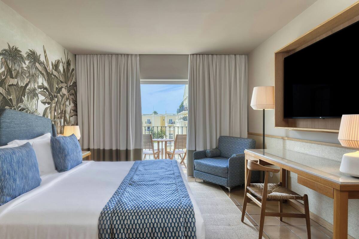 A hotel room with a large bed, a desk, a chair, a TV, and a balcony with a view of buildings.