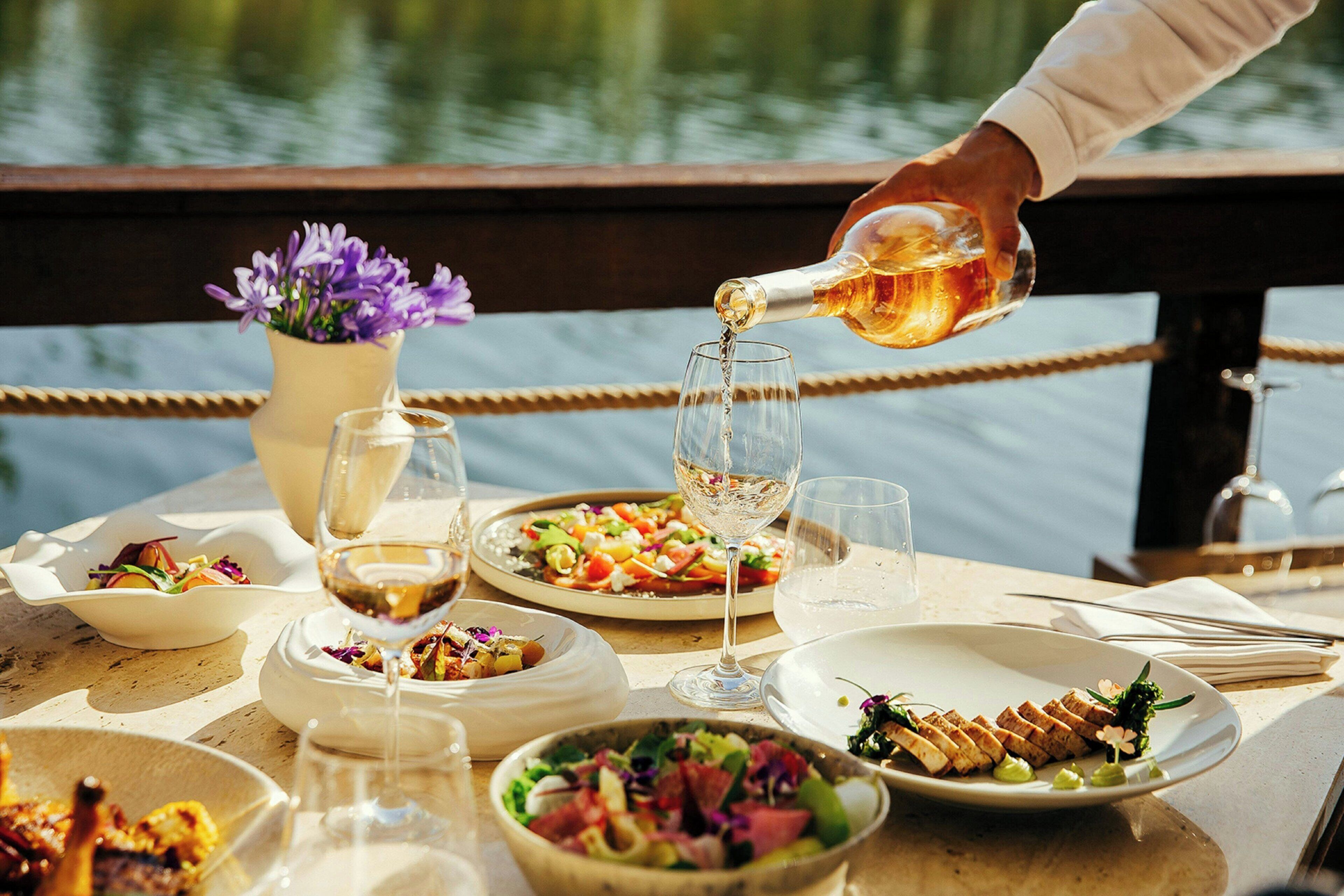 A table set with various dishes and glasses of wine, with a body of water in the background.