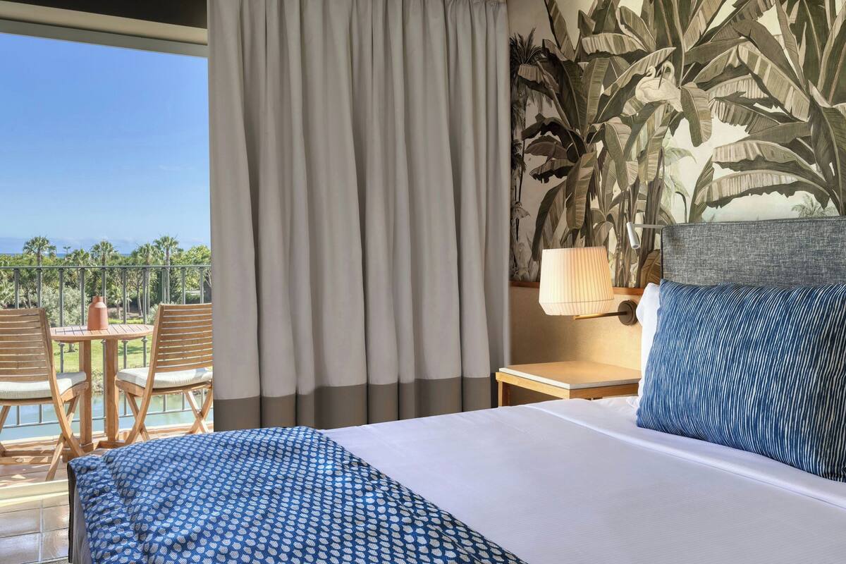 A hotel room with a bed, a nightstand, a lamp, and a view of a balcony with chairs and a table.