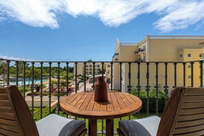 A balcony with a wooden table and two chairs, overlooking a pool and a building with balconies.
