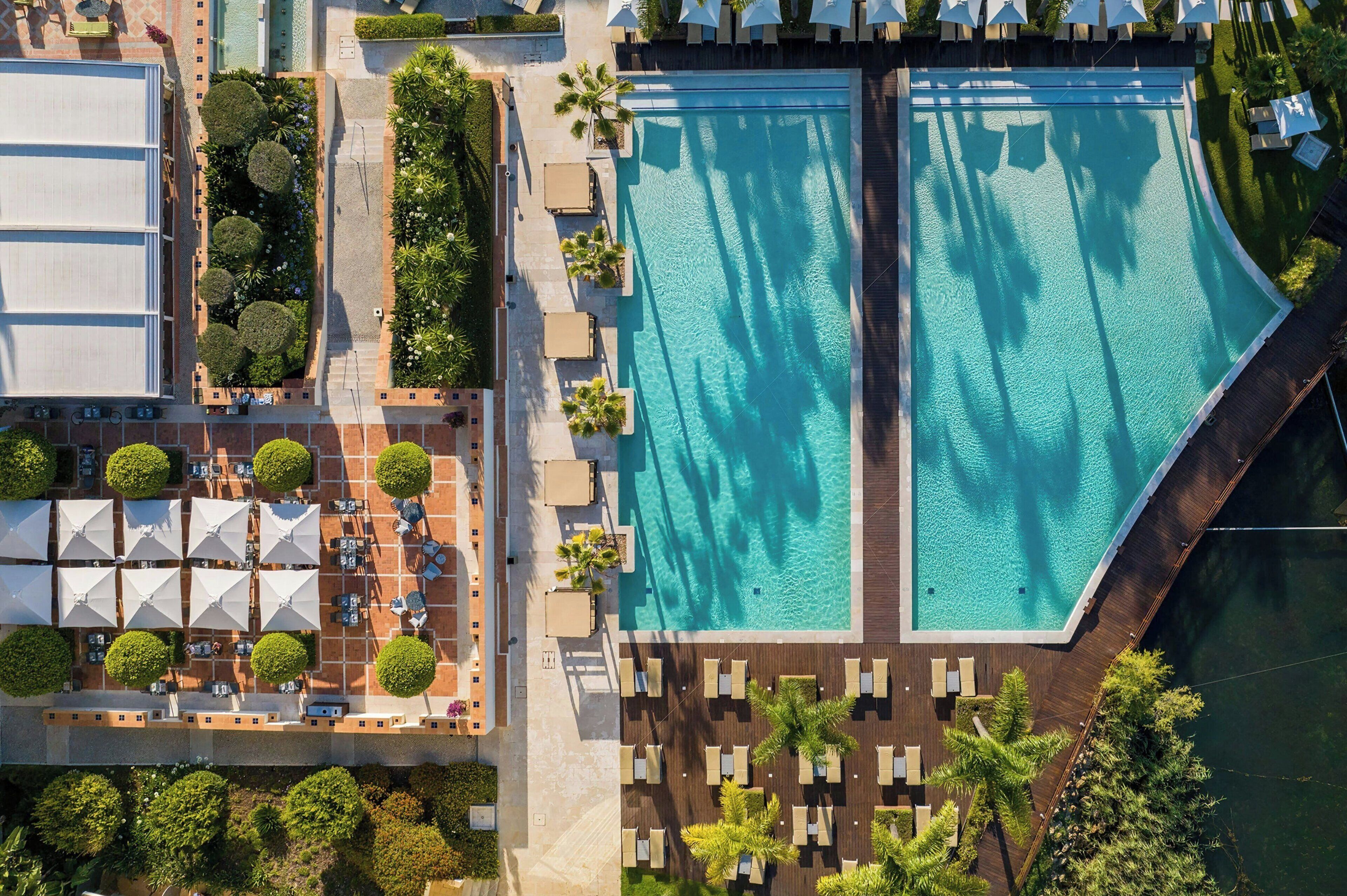 An aerial view of a resort with two large swimming pools, sun loungers, umbrellas, and palm trees.