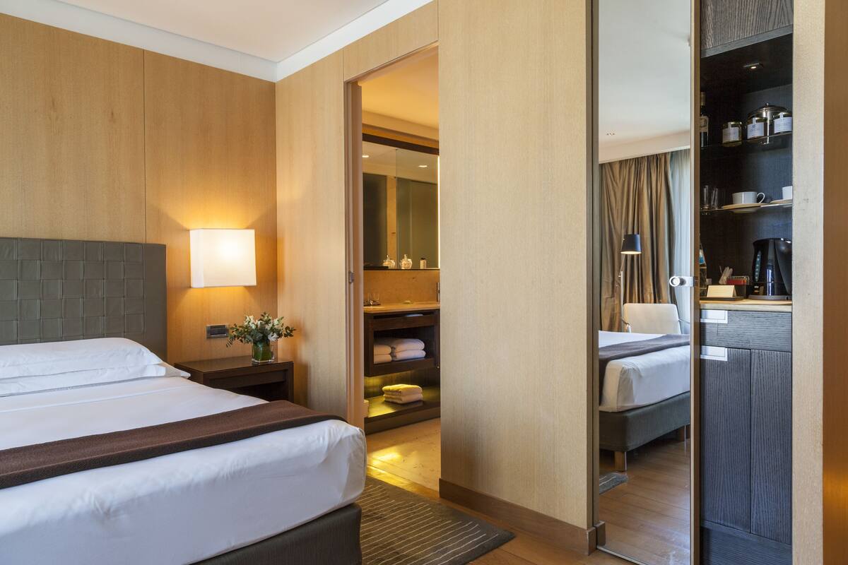 Une chambre d’hôtel avec deux lits, une table de chevet avec une lampe, un miroir, une armoire et une salle de bain visible à travers une porte ouverte.