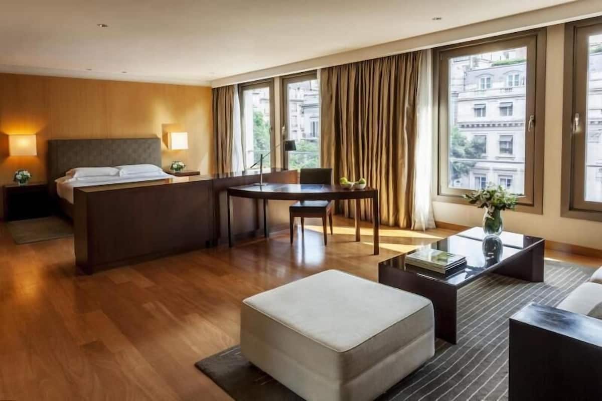 Une chambre d’hôtel comprenant un lit, un bureau, une table à manger, une table basse et de grandes fenêtres avec des rideaux.