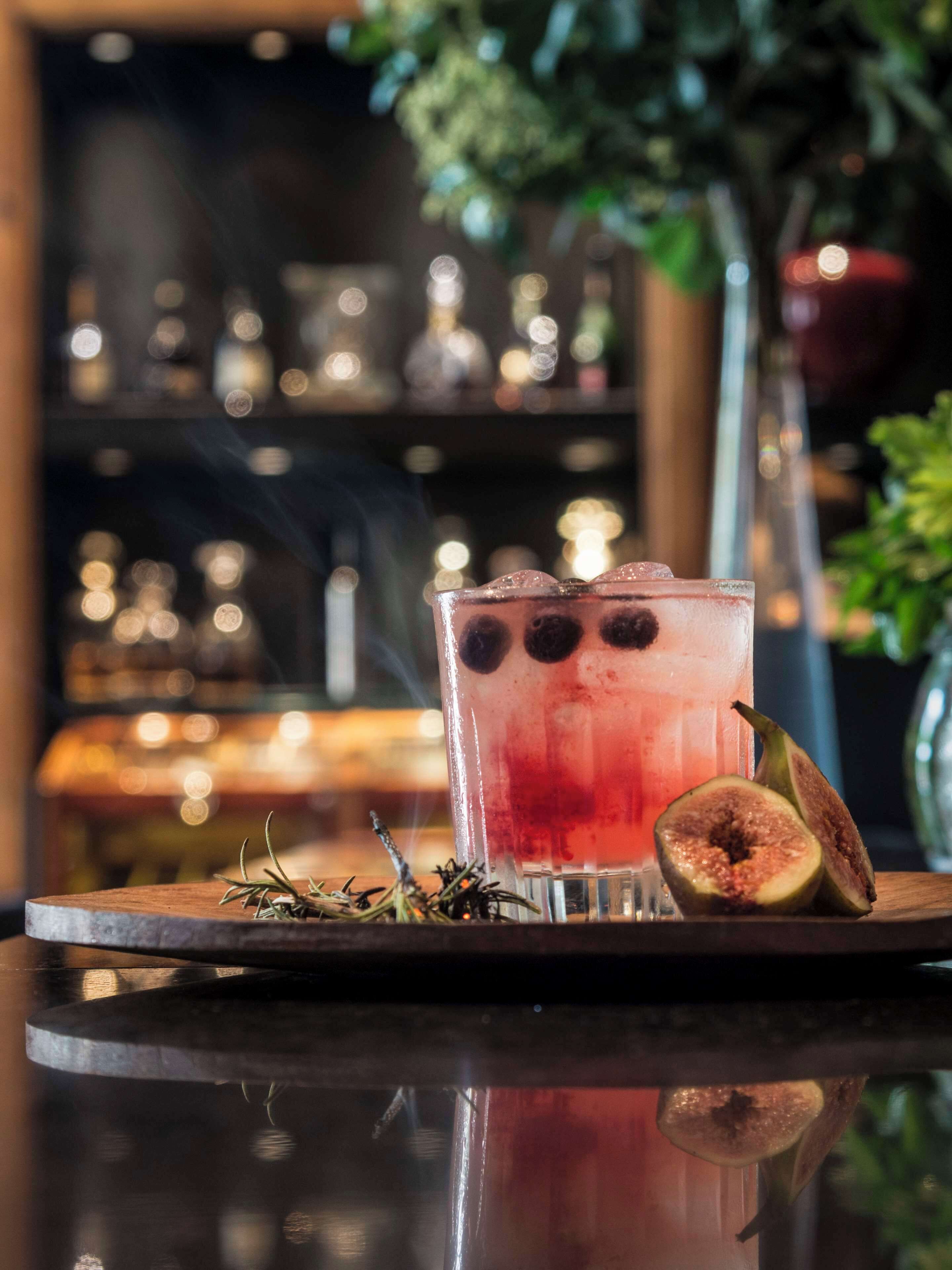 A glass of red beverage with ice cubes and a halved fig on a wooden coaster, with a blurred background of a bar setting.