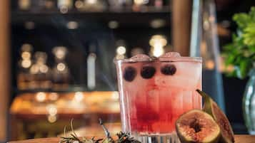 A glass of red beverage with ice cubes and a halved fig on a wooden coaster, with a blurred background of a bar setting.