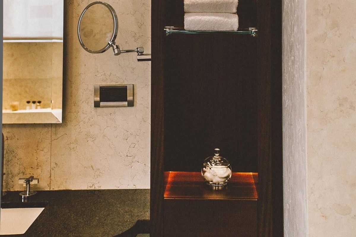 Une salle de bain avec un miroir, deux lampes fixées au mur, une œuvre d’art encadrée, un porte-serviettes avec des serviettes pliées, un savonnière en verre et un porte-rouleau de papier toilette.