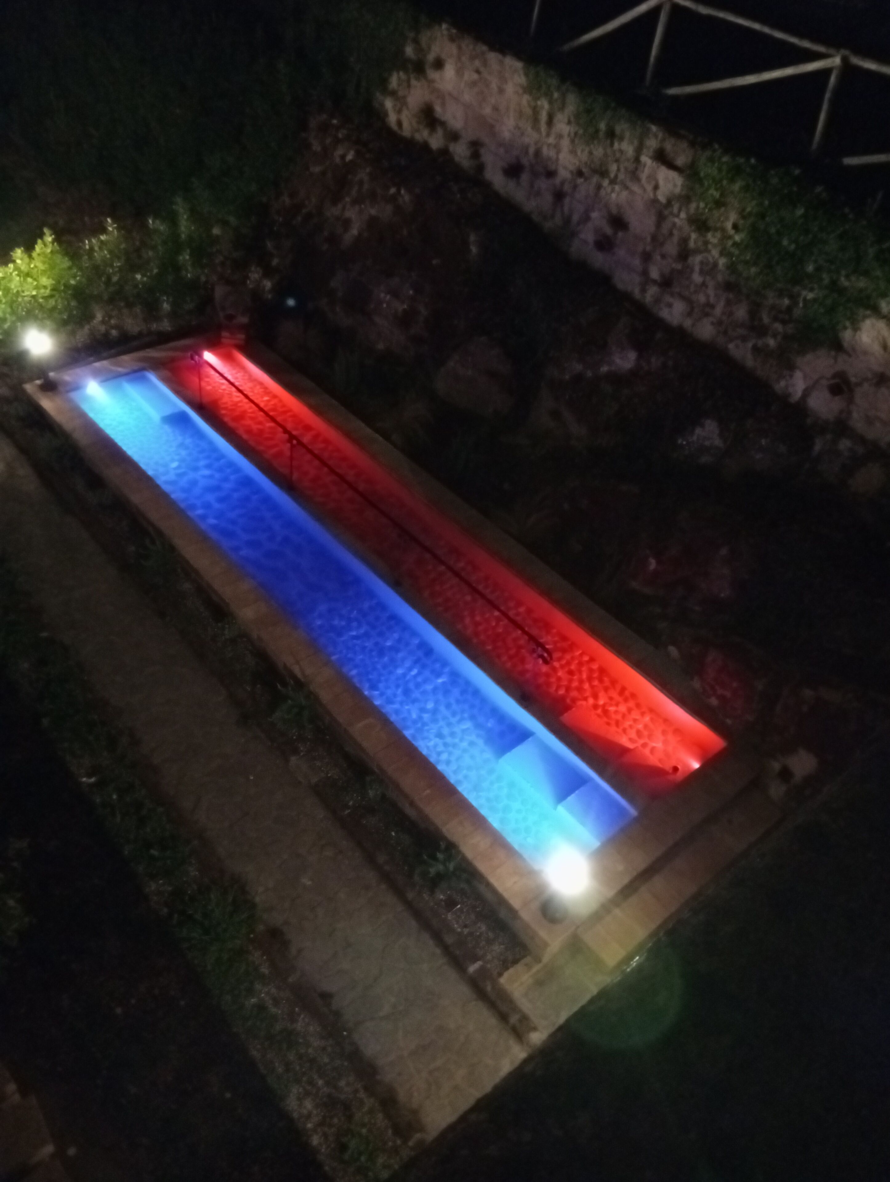 Una piscina rettangolare con bordo illuminato in blu e rosso, circondata da un muro in pietra e vegetazione.