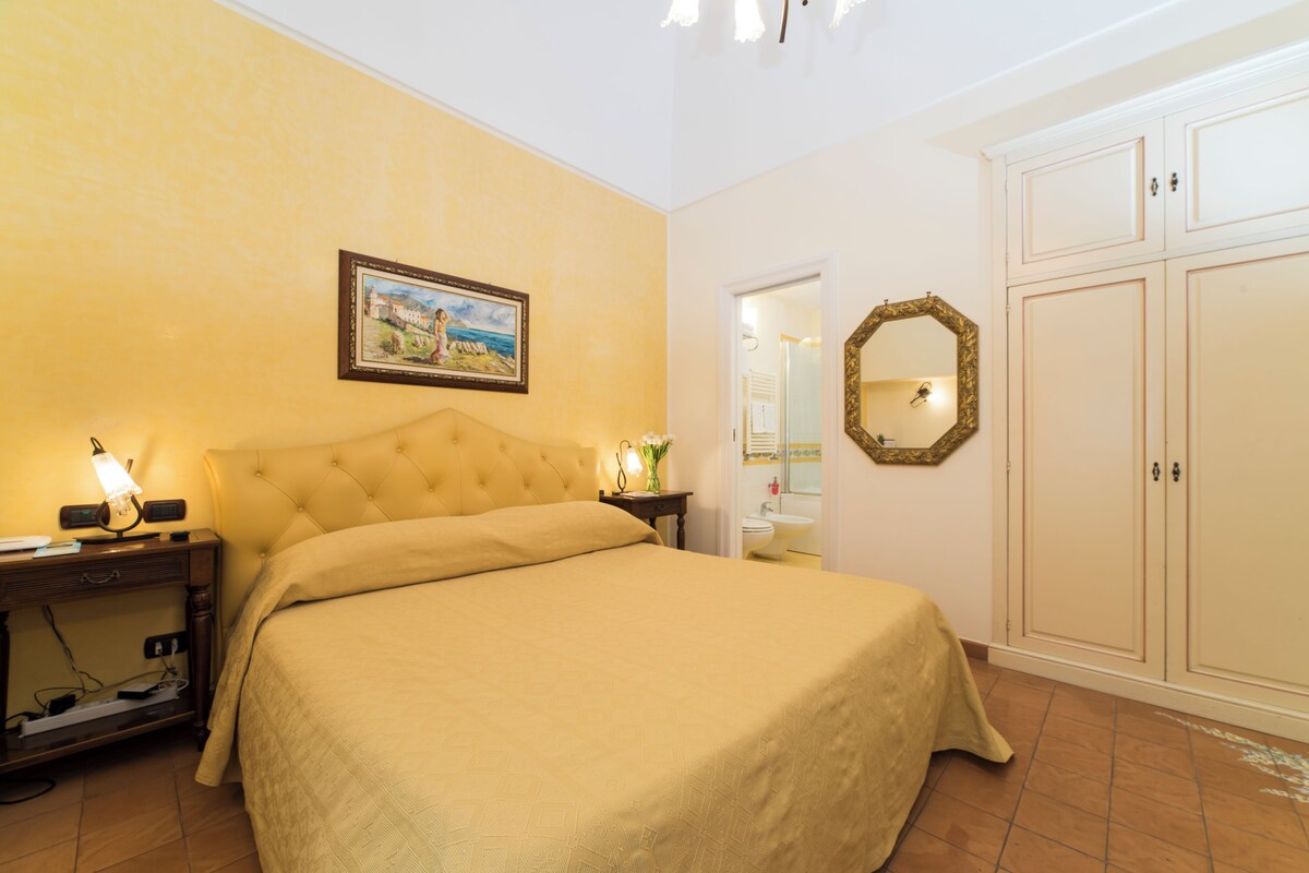 A bedroom with a bed, bedside lamps, a nightstand, a framed picture on the wall, a mirror, and a wardrobe.