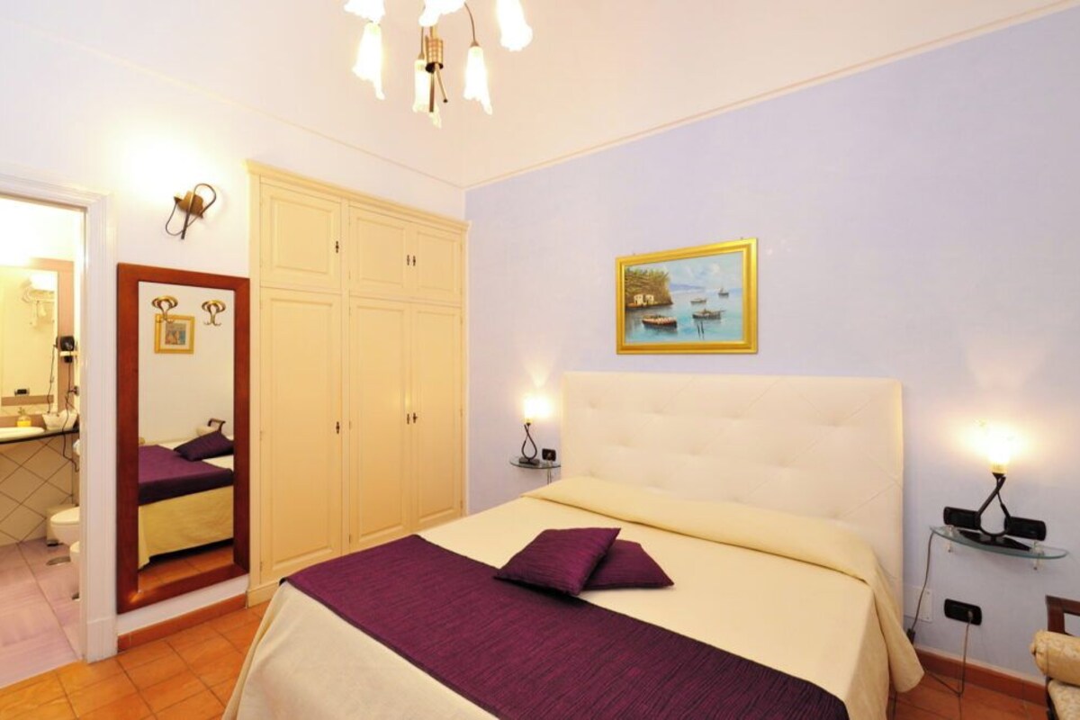 Een slaapkamer met een bed, nachtkastje, spiegel, kledingkast en een schilderij aan de muur.