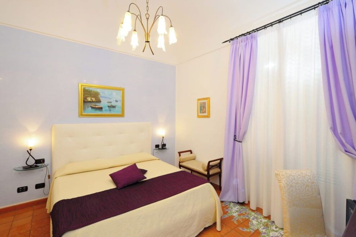 Ein Schlafzimmer mit einem Bett, Nachttischlampen, einem Sessel, einem kleinen Tisch und einem Fenster mit Vorhängen.