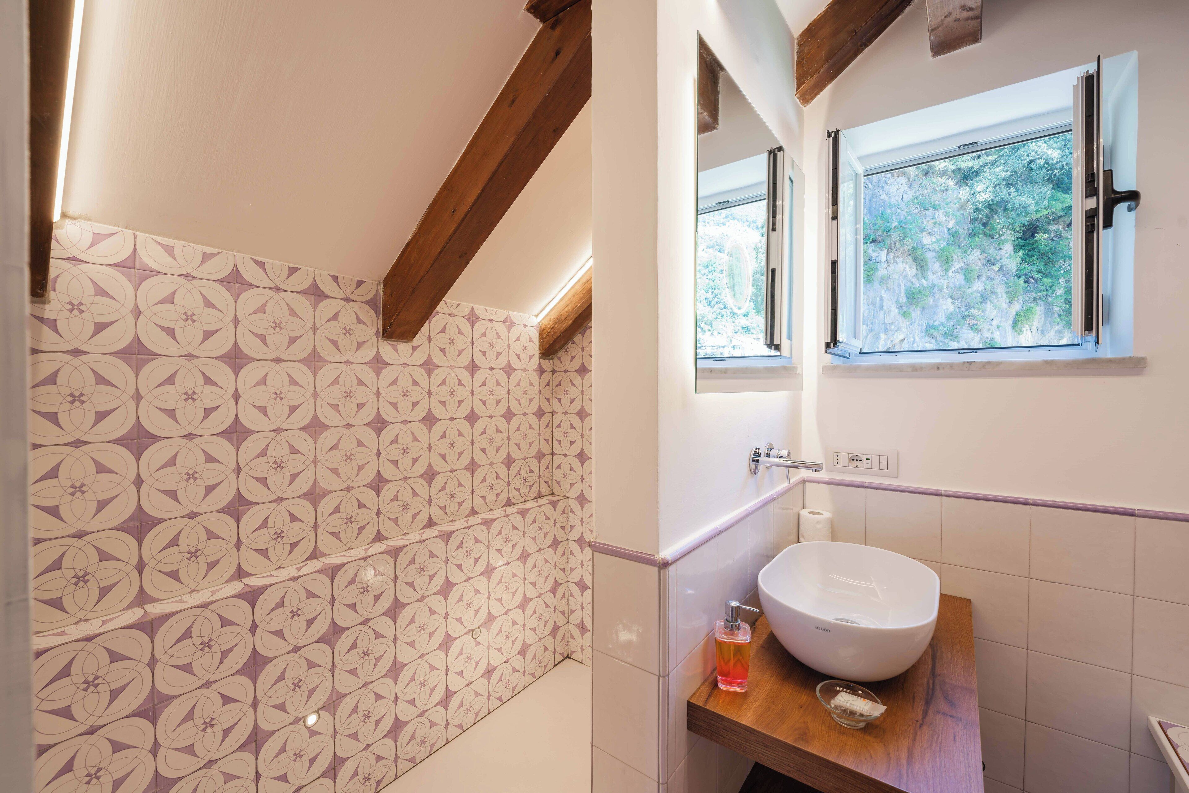 Ein Badezimmer mit gemusterten Fliesen, Holzbalkendecke, großem Fenster und einem weißen Waschbecken.