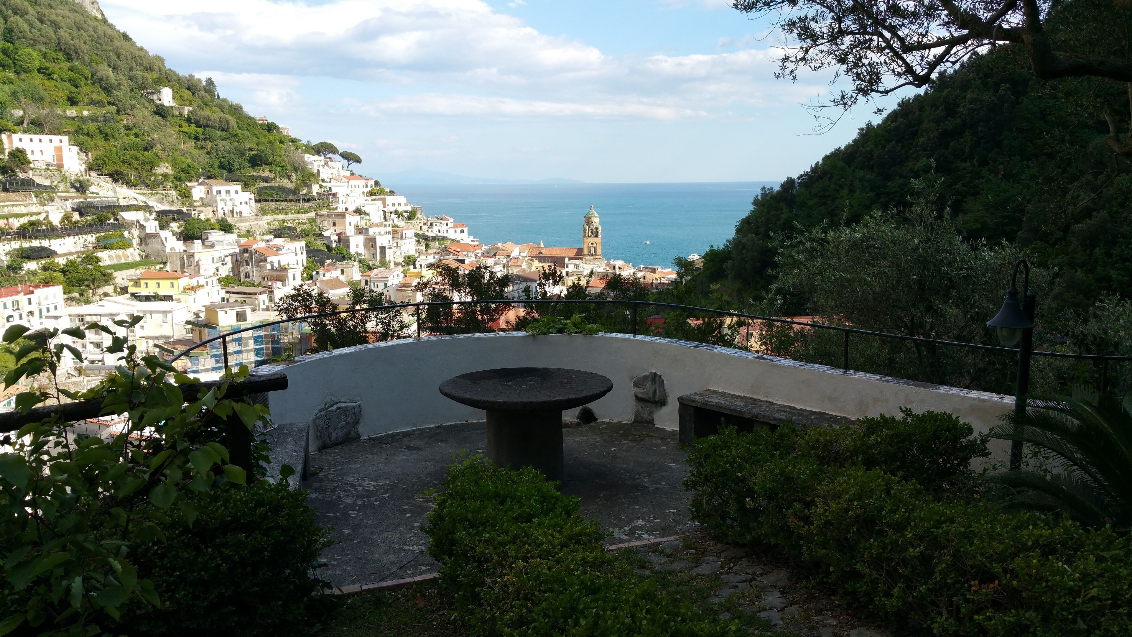 A terrace with a stone table and benches, overlooking a coastal town with buildings and a church.