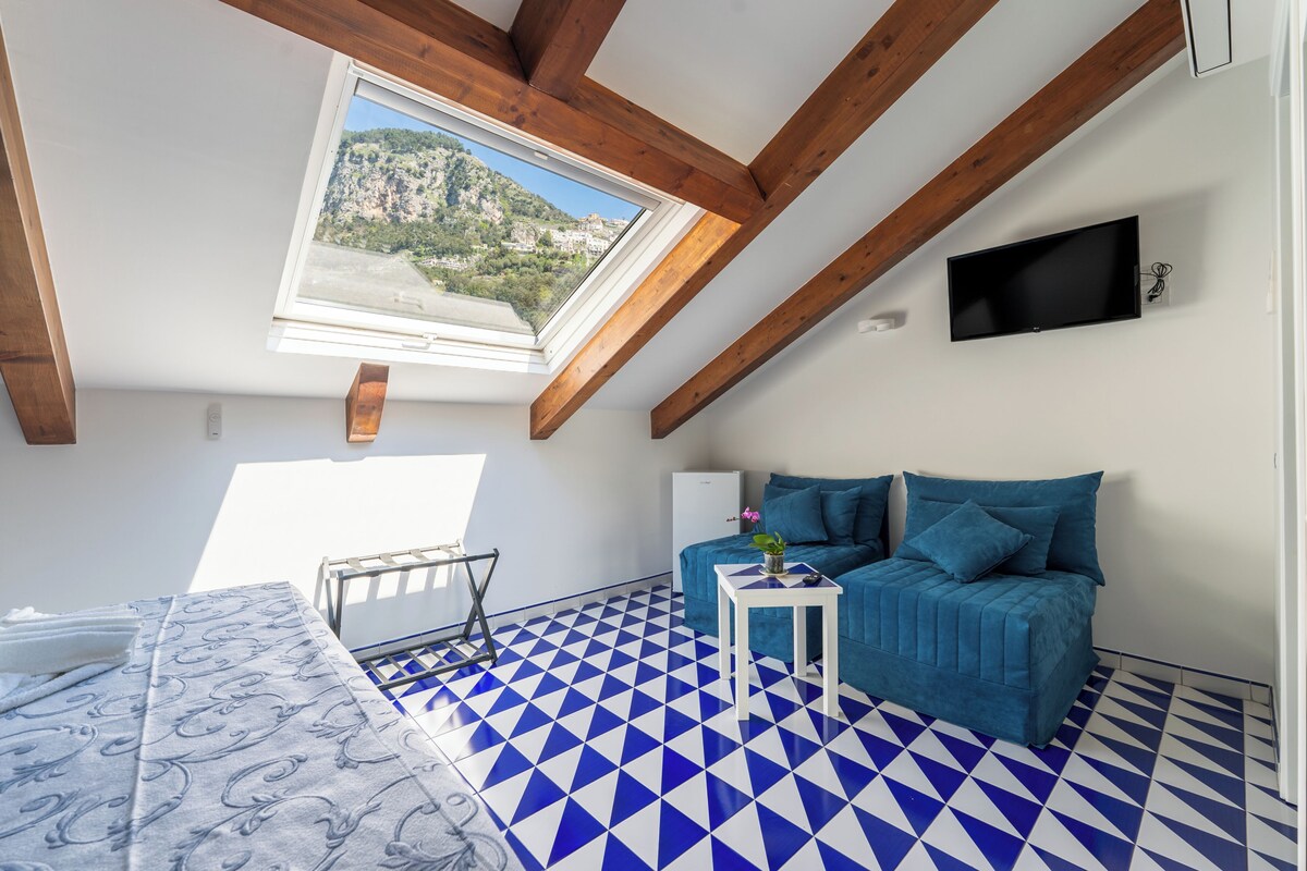 Ein Zimmer mit blau-weiß kariertem Boden, einem blauen Sofa, einem kleinen weißen Tisch, einem an der Wand befestigten Fernseher und einem Fenster mit Blick auf eine Berglandschaft.