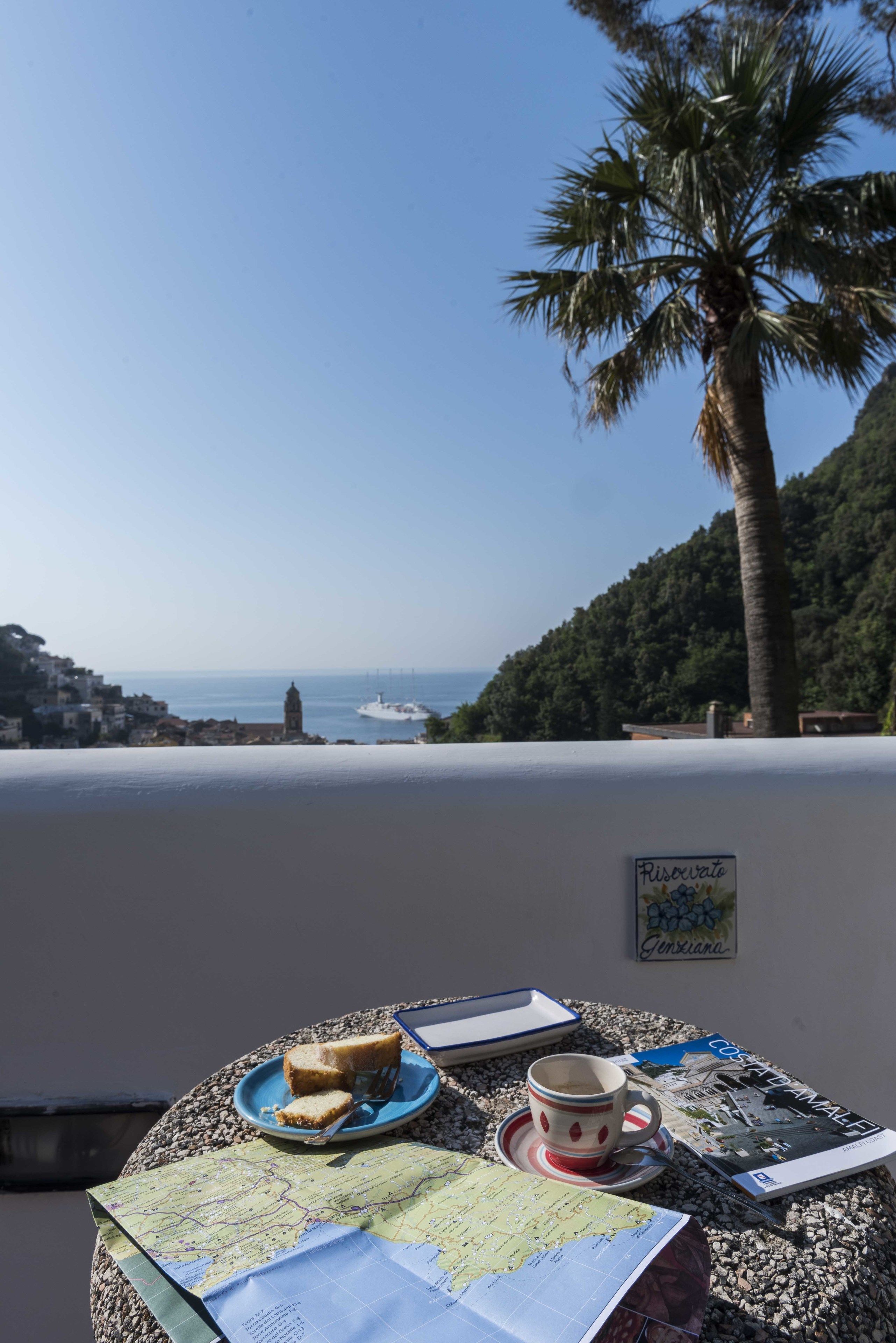 Ein Tisch mit einer Karte, einem Teller mit Brot, einer Tasse und einem Buch, mit Blick auf eine Küstenlandschaft mit einer Palme und Gebäuden.